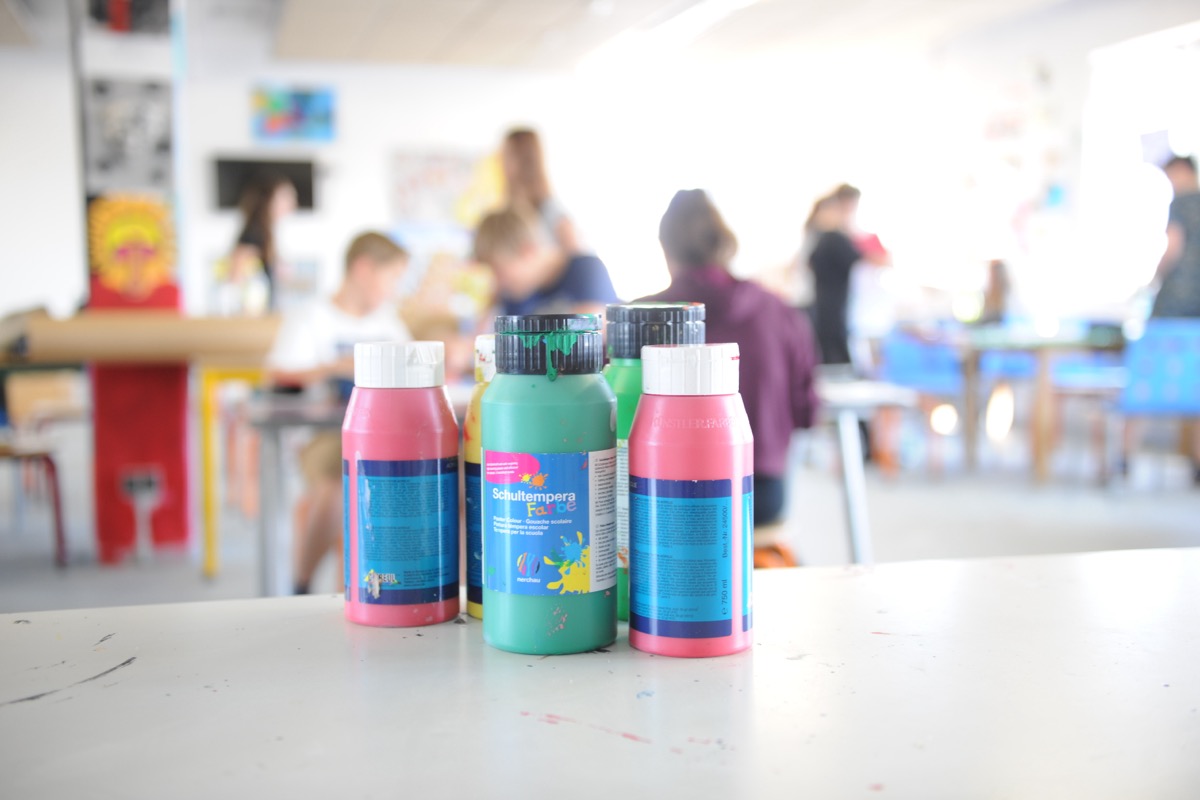 Bunte Farbflaschen auf einem Tisch, im Hintergrund malen Kinder